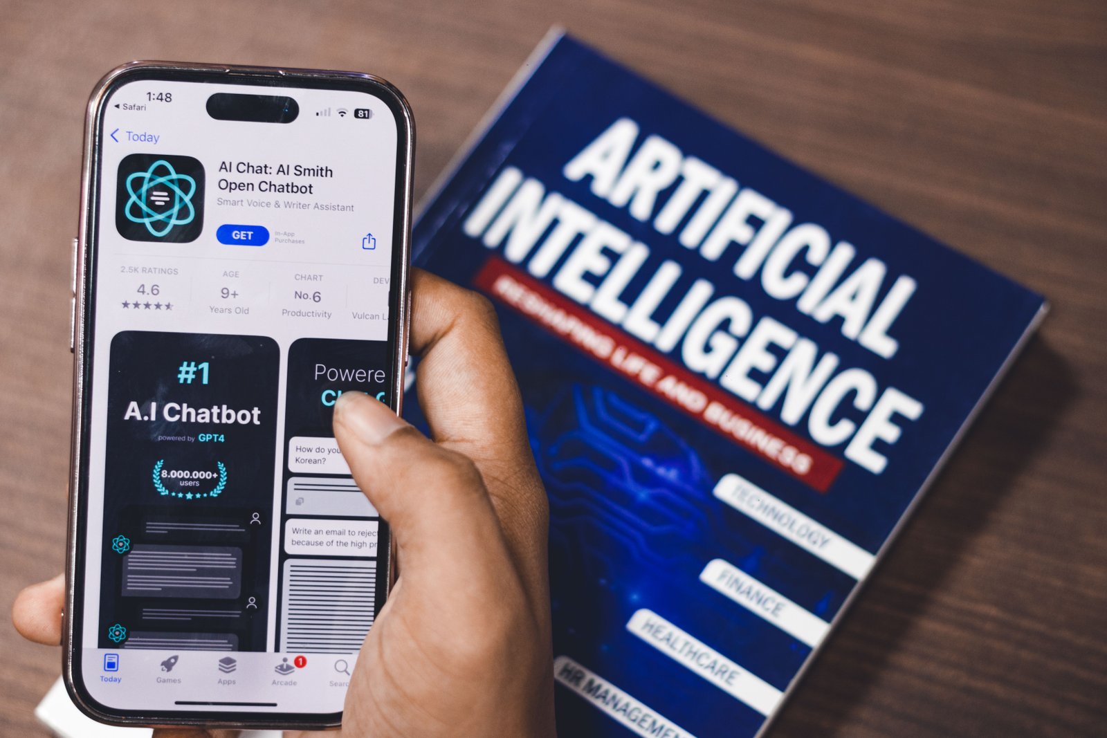 A hand holding a smaetphone showing an AI chatbot app next to an Artificial Intelligence book on a desk